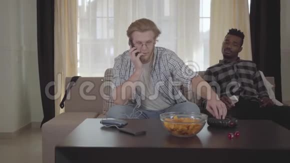 一个英俊的长胡子男人坐在沙发上用手机说话而他的非裔美国人朋友正看着他的背影视频的预览图