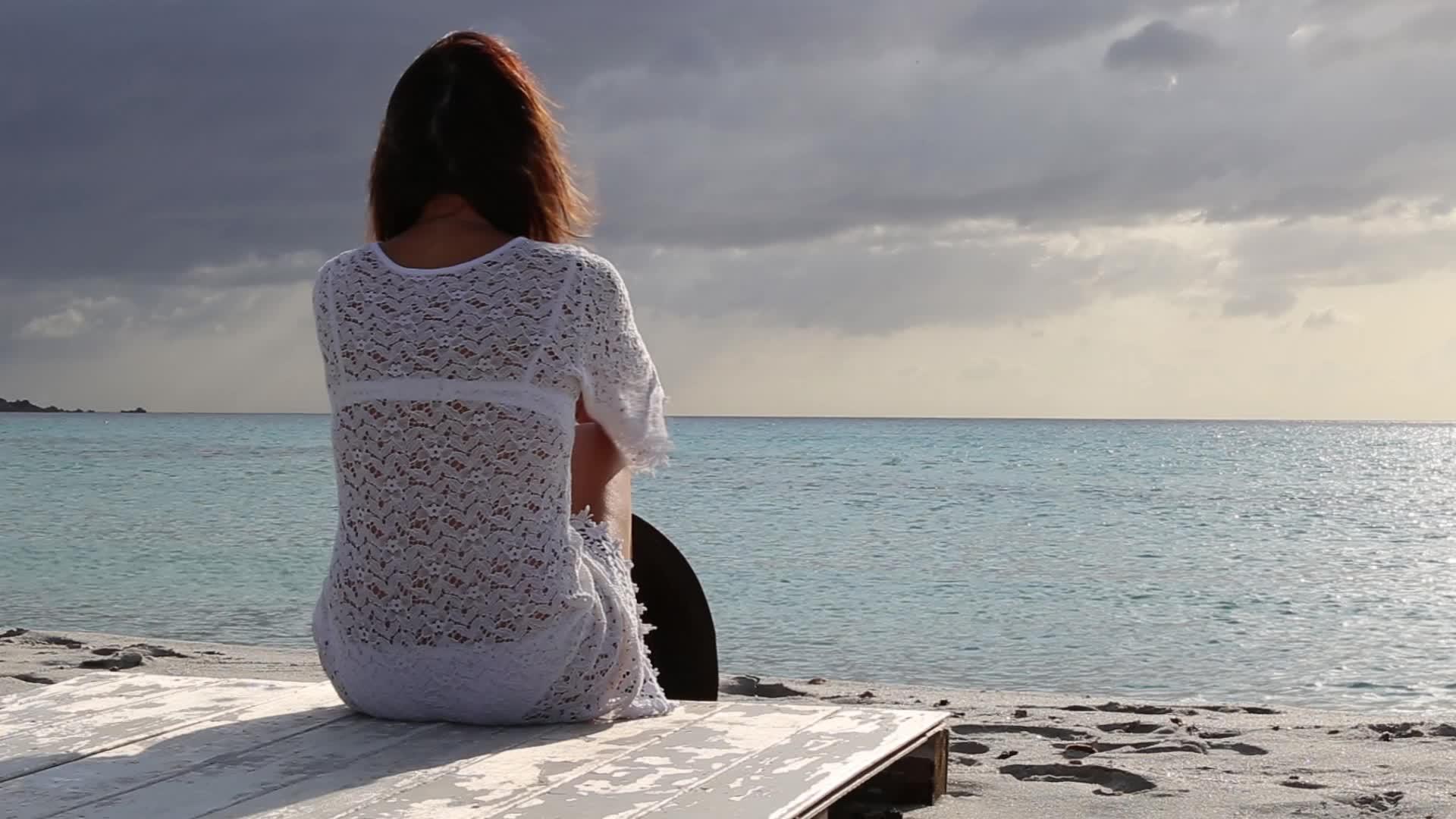 年轻女性坐在海边在黎明风中看地平线视频的预览图