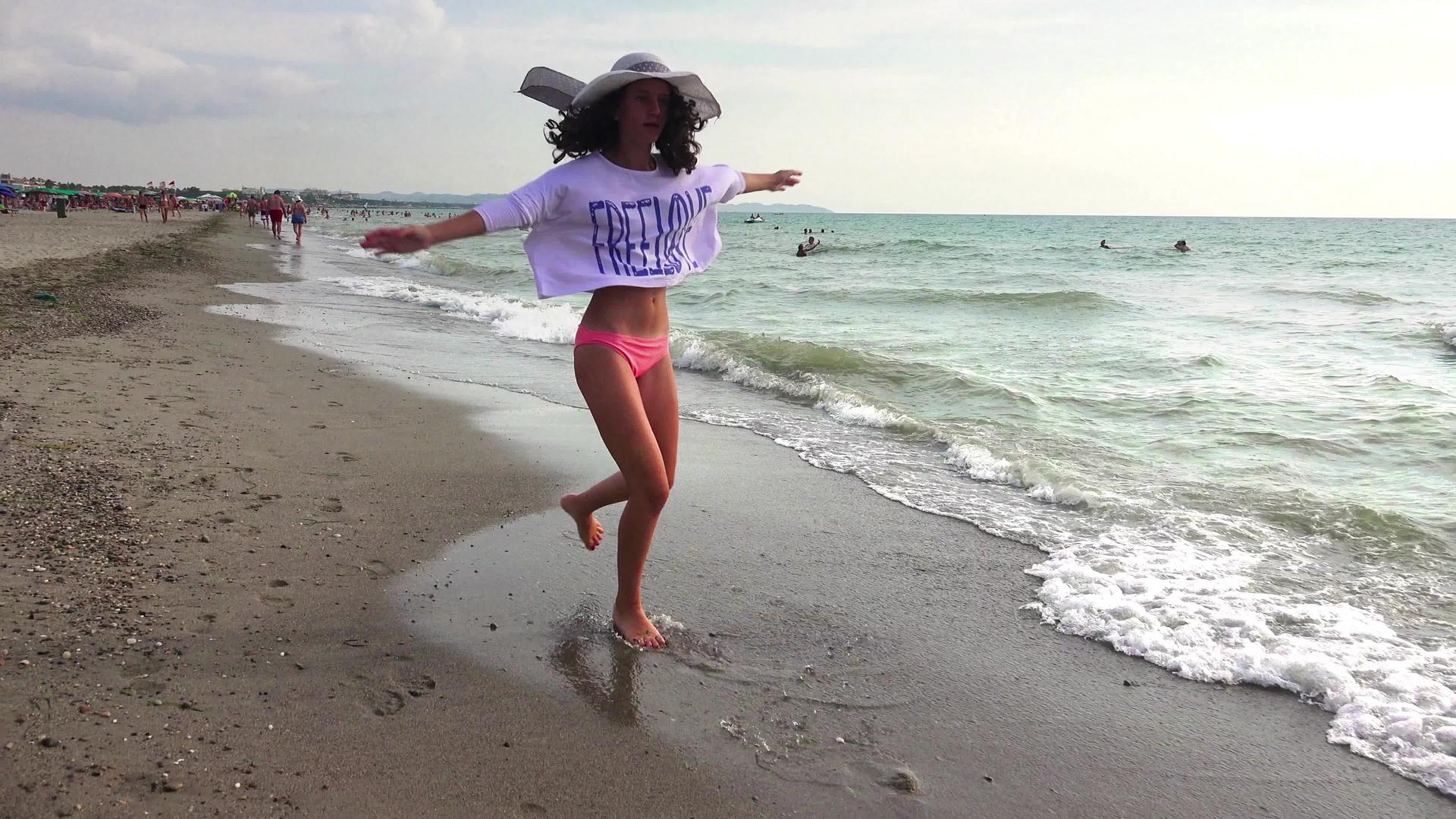 戴着帽子的漂亮女孩享受暑假快乐地跑步跳上海滩冲向海浪视频的预览图