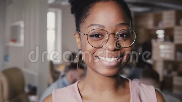 年轻漂亮的非裔美国人戴着眼镜的快乐女领导在现代办公室工作背景4K对着镜头微笑视频的预览图