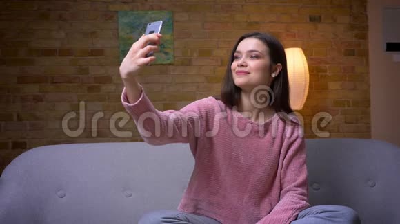一位年轻漂亮的黑发白种人女性在电话上自拍坐在沙发上微笑的特写镜头视频的预览图