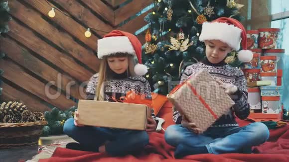 小双胞胎坐在圣诞树下摇晃着摆弄着礼物盒找出里面是什么视频的预览图