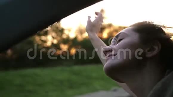 漂亮的女人坐在车里幸福地闭上眼睛手在空中玩耍太阳落山了风吹头发慢节奏视频的预览图
