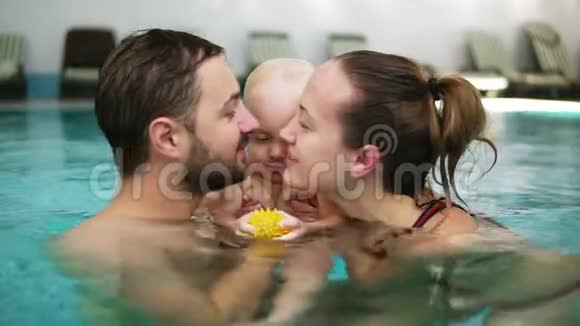 年轻的母亲和父亲正在游泳池里抱着他们可爱的孩子在他们的嘴唇上亲吻对方快乐视频的预览图