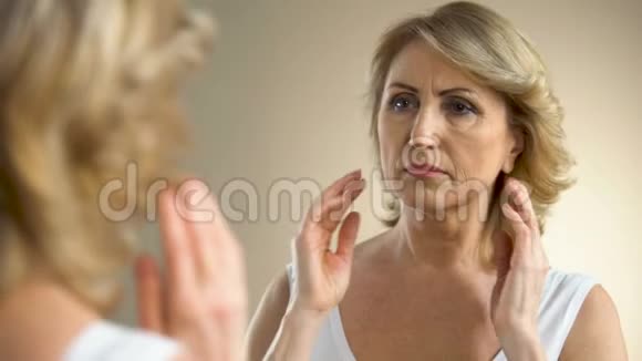 不高兴的老妇人在家照镜子摸她的脸衰老的过程视频的预览图