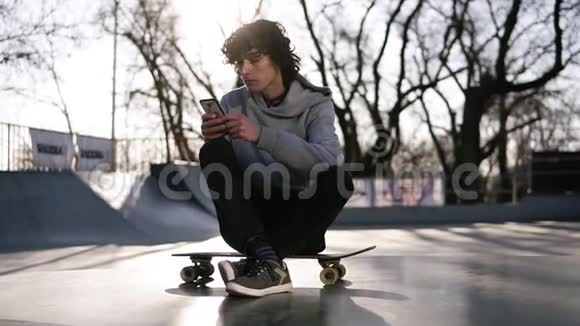 年轻人正拿着手机坐在滑板公园的溜冰区一个人正在聪明的人上网视频的预览图
