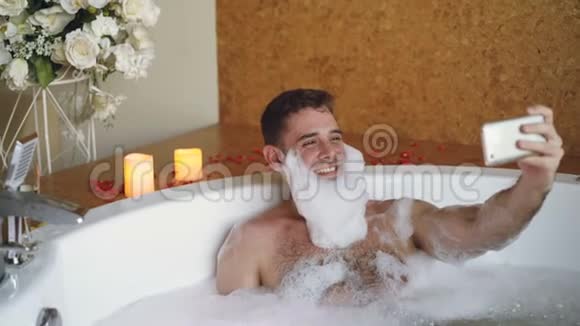 在现代水疗沙龙里带泡沫胡须的开朗男人正在用智能手机在热水浴缸里自拍他在笑视频的预览图