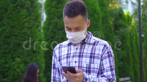 戴着医用口罩的亚洲年轻人在公园里咳嗽用智能手机慢吞吞地说视频的预览图