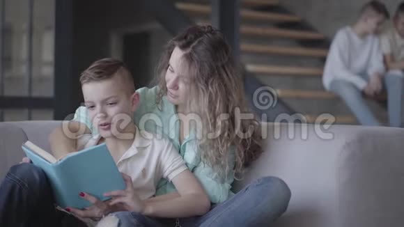年轻漂亮的母亲和儿子坐在沙发上看书的画像而她的两个孩子则坐在沙发上视频的预览图