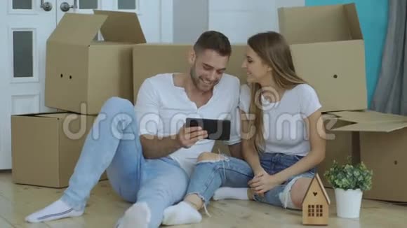 年轻的幸福夫妇坐在地板上用平板电脑在他们的新家新家里找到重新煎煮的交通服务视频的预览图