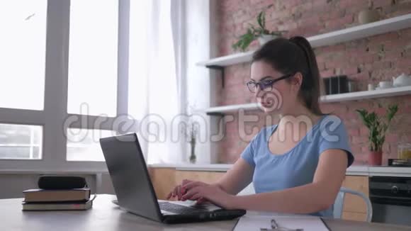 在线培训戴眼镜的英俊年轻女子使用笔记本电脑并在坐在厨房办公桌前的剪贴板上写笔记视频的预览图