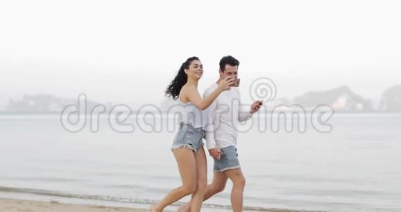 情侣在沙滩上行走使用手机智能手机牵手年轻男子女子在网上旅游视频的预览图