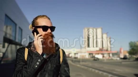 在城市大街上戴着墨镜笑容可掬会说话的智能手机的年轻留胡子的嬉皮士多莉拍摄视频的预览图