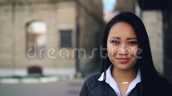 一幅迷人的亚洲女孩的特写慢镜头微笑着看着镜头站在街上穿着视频的预览图