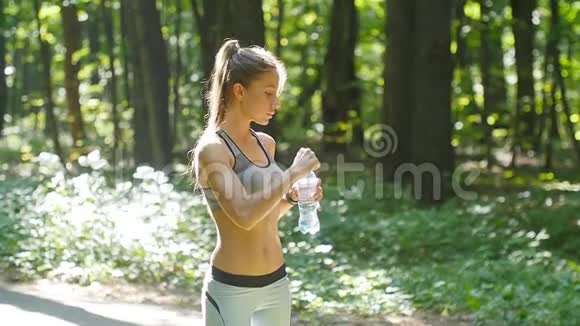健身妇女从阳光明媚的森林里的瓶子里喝水视频的预览图