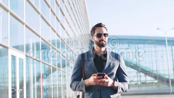 时尚留胡子的男士在经过机场候机楼时戴着太阳镜对着收到的短信微笑视频的预览图