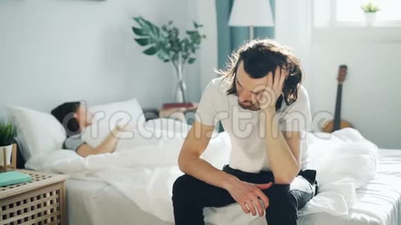 不快乐的丈夫坐在床上在妻子睡在家时摸着悲伤的脸视频的预览图