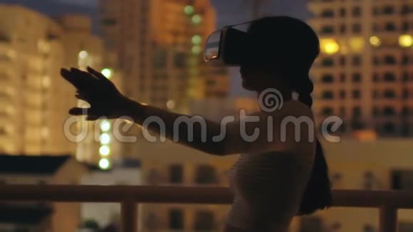 使用虚拟现实耳机在屋顶露台上缓慢移动年轻女性晚上有VR体验视频的预览图