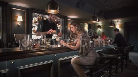 当年轻的沮丧的女人坐在酒吧柜台附近时Hipster酒保把配料和鸡尾酒结合在酒吧里视频的预览图