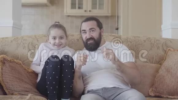 大胡子男人坐在沙发上看电视他的小女儿支持足球队关门女孩跳起来视频的预览图