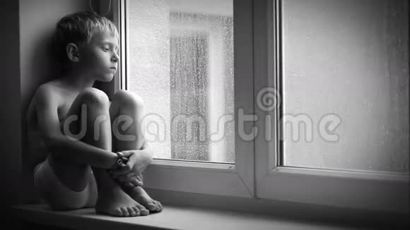一个悲伤的男孩在倾盆大雨中坐在窗台上无法走出公寓的黑白镜头视频的预览图