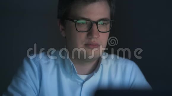 戴眼镜的年轻人晚上在黑暗的房间里用笔记本电脑工作视频的预览图