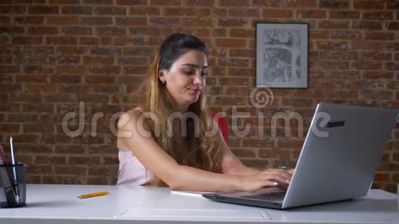 疲惫的工作妇女使用她的笔记本电脑一边打字一边坐在桌面上的砖墙上无聊的脸视频的预览图