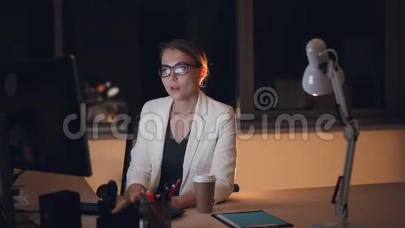 戴着眼镜和西装的疲惫的金发女郎深夜在电脑上工作喝着外卖咖啡感到疲倦和疲惫视频的预览图