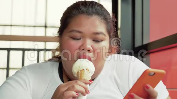 肥胖的女人吃冰淇淋用手机视频的预览图