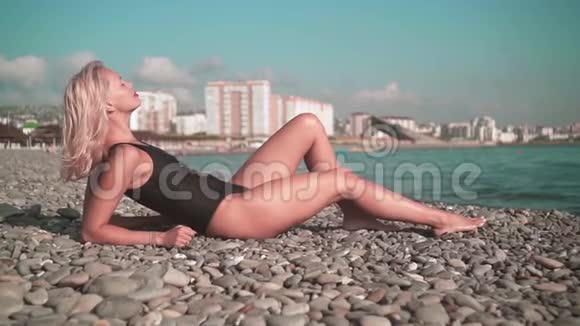 穿着泳衣躺在岩石海岸上的年轻性感女孩金发碧眼的一件泳衣躺在海滩的石头上视频的预览图