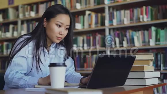 年轻的亚洲学生女孩坐在桌子旁大学图书馆里有一堆书用手提电脑工作喝酒视频的预览图
