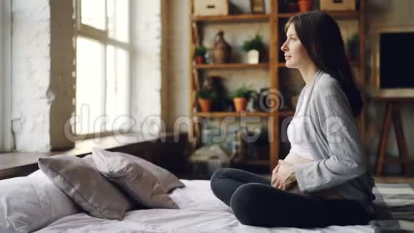 可爱的怀孕女孩抚摸着她的腹部躺在现代的平躺在床上看着窗外然后闭上眼睛视频的预览图