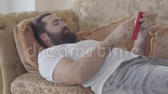 英俊的胡子男人躺在沙发上检查手机他生气了扔掉了智能手机接收坏消息视频的预览图