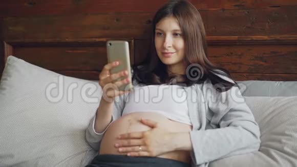 美丽的怀孕少女黑发正在抚摸她的婴儿肿块并使用智能手机在家里的床上轻松愉快的微笑视频的预览图