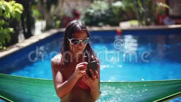美丽的年轻黑发女人戴着太阳镜比基尼坐在吊床上用手机在游泳池里自拍视频的预览图