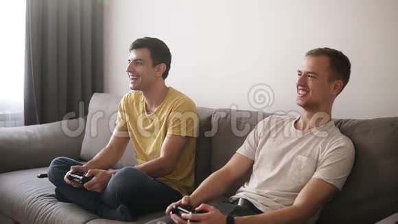 两个年轻人在家玩电子游戏拿着操纵杆坐在阁楼内部房间的灰色沙发上微笑视频的预览图