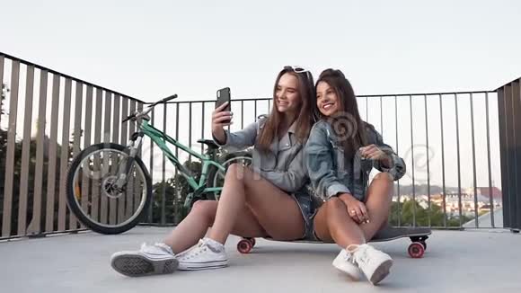 两位女性最好的朋友在夏日阳光明媚的日子里坐在滑板上用智能手机自拍视频的预览图