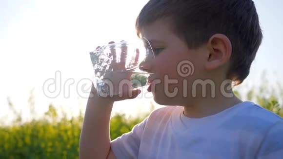 快乐的小男孩在背景花上喝干净的水小男孩在户外喝玻璃孩子视频的预览图