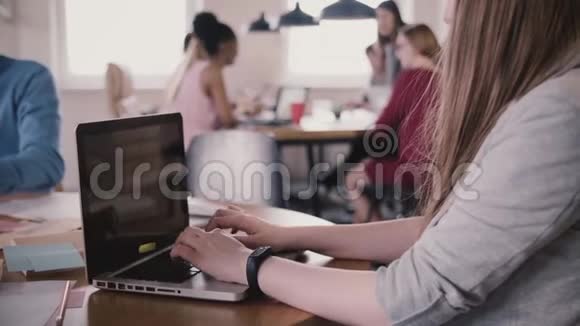 年轻的自由职业女性员工在忙碌的现代工作空间里在桌旁用智能手表在笔记本电脑上打字视频的预览图
