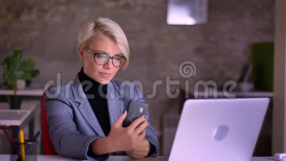 戴眼镜的中年金发短发女商人在办公室用手机制作自拍照片的肖像视频的预览图