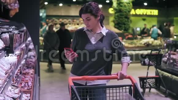 年轻的深色头发的女顾客在杂货店里穿过肉道时看着手机女孩带着腊肠视频的预览图