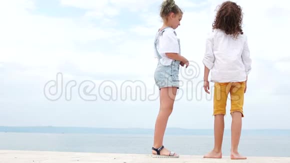 女孩和男孩站在一个海洋泊位上女孩脱下凉鞋赤脚走暑假快乐的孩子们视频的预览图
