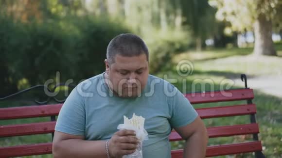 一个胖子在长凳上吃东西看着他有害可口的快餐视频的预览图