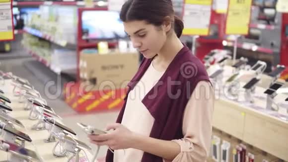 年轻漂亮的黑发女孩在电子商店摘电话时微笑查看智能手机应用程序视频的预览图
