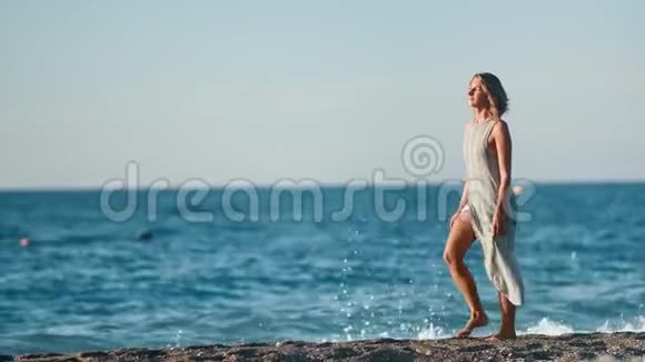 迷人的晒黑旅游女人穿着连衣裙在日落时分在海滩上散步全镜头视频的预览图