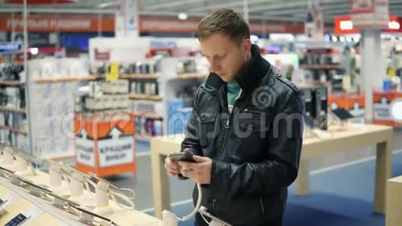 穿黑色皮夹克的年轻人正在商店里挑选一部新手机检查它的工作原理视频的预览图