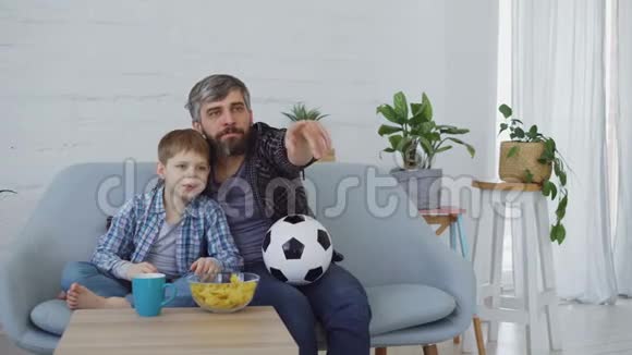 家庭成员父子俩在家里看足球比赛欢呼庆祝胜利吃零食视频的预览图