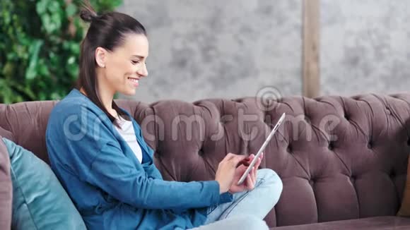 侧视笑少女使用平板电脑坐在舒适客厅的沙发上视频的预览图