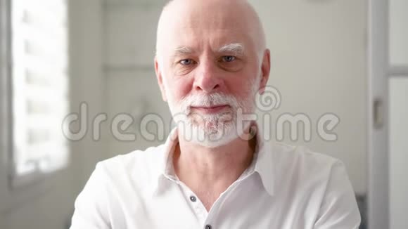 英俊的留胡子老人在家的肖像坐在客厅里看着摄像机视频的预览图