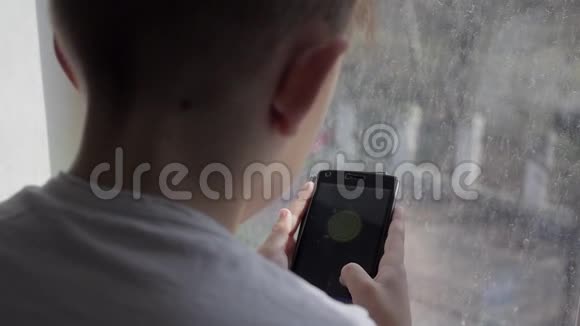 小男孩在家里玩智能手机游戏雨天男孩坐在窗台上视频的预览图
